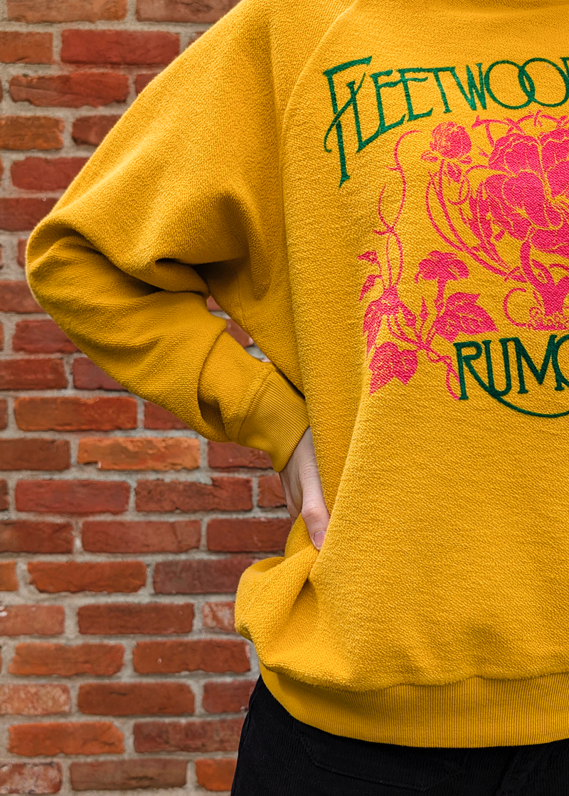 Close up details of the Fleetwood Mac Rumours sweatshirt with a reversed inside out textured look. Features a crew neckline, raglan sleeves, and ribbed cuffs, neckline, and hem. Golden Daze mustard colorway with flocked velvet writing in green and pink floral details. 100% French terry, made in the USA